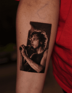 Tattoo of a musician holding a microphone on a forearm, showcasing black and grey realism style, reflecting Pony Lawson's artistic focus on detailed portrait tattoos.